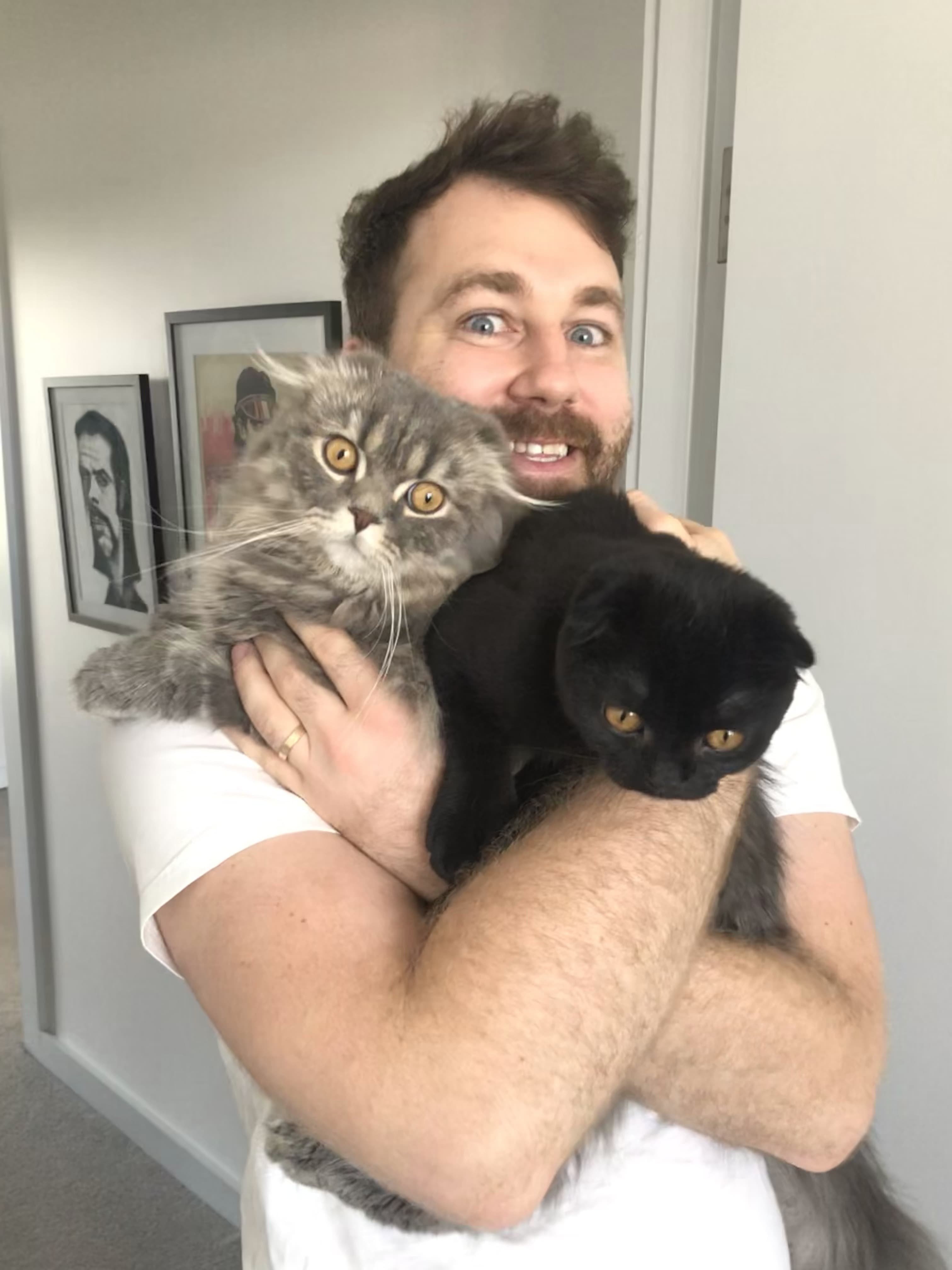 Greg holding his two cats Sesame and Rame and smiling excitedly